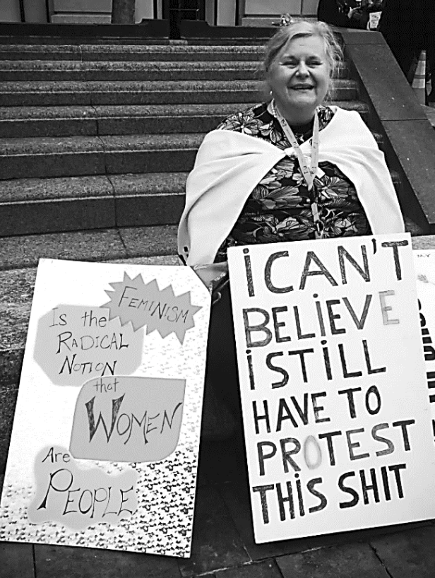 Person auf Treppe mit Protestschildern: "I can't believe I still have to protest this shit" und feministische Botschaft.